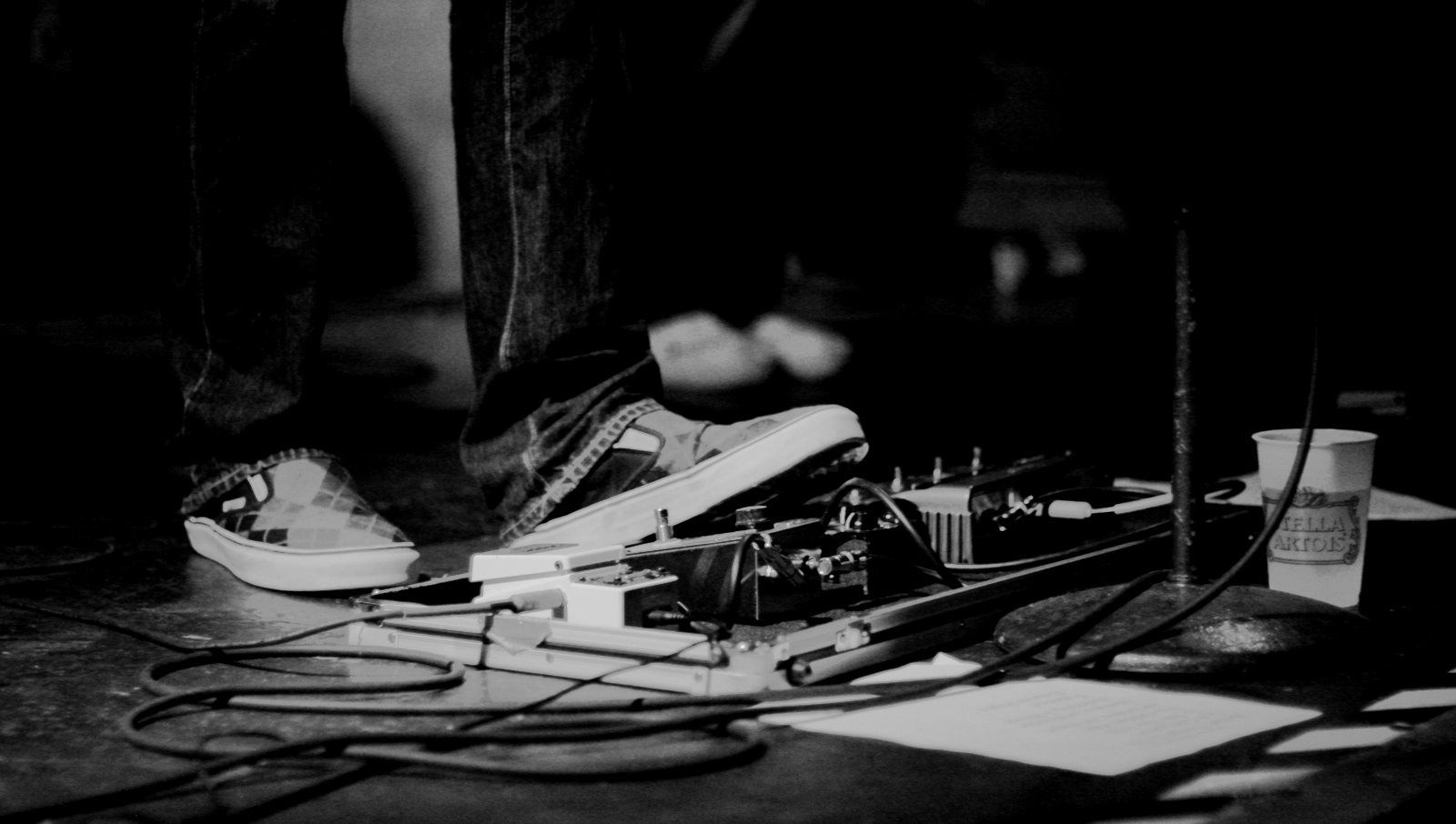 A closeup photograph of a guitar effect pedalboard onstage, with the lower third of the user's legs visible.